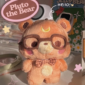 Peut inclure: Un porte-clés en peluche en forme d'ours de couleur pêche, portant des lunettes à monture marron et un nœud papillon. L'ours a un croissant de lune sur le front. Le texte sur l'image indique "meet Pluto the Bear" et "Plushie Keychain".