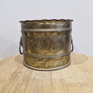 May include: A small, aged metal bucket with a decorative, scalloped rim. The bucket has two small handles and a tarnished, brassy finish. The bucket is sitting on a wooden surface.
