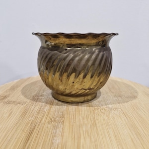 May include: A small, antique brass planter with a wavy rim and a spiral design. The planter has a round base and a slightly flared opening. The brass has a warm, golden hue with some areas of tarnish, giving it a vintage appearance.