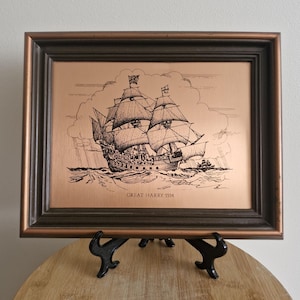 May include: A framed copper-coloured print of a sailing ship, with the text "GREAT HARRY 1514" at the bottom. The print is in a dark brown frame, and is displayed on a black stand. The ship is depicted sailing on rough seas.