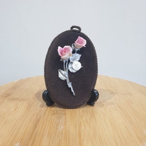 May include: A small oval-shaped framed picture with a brown velvet background. A silver-coloured rose with pink porcelain flowers is displayed in the centre of the frame.