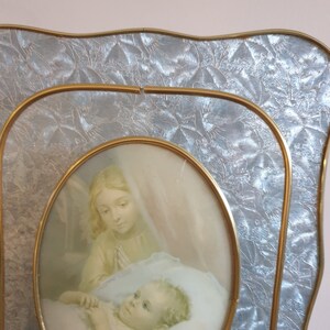 Op de afbeelding: Een gouden fotolijst met een reli&euml;f, textuur, zilverkleurige achtergrond. De lijst bevat een afbeelding van een vrouw in een witte jurk die een slapende baby vasthoudt.