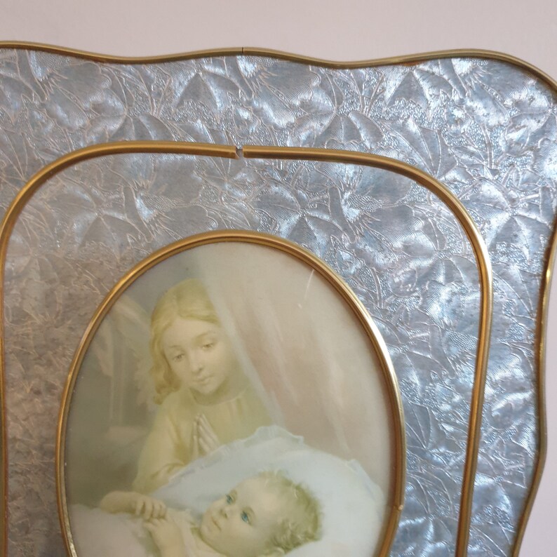 Op de afbeelding: Een gouden fotolijst met een reli&euml;f, textuur, zilverkleurige achtergrond. De lijst bevat een afbeelding van een vrouw in een witte jurk die een slapende baby vasthoudt.