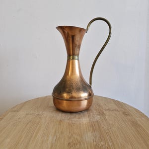 May include: A copper jug with a brass handle. The jug has a bulbous body and a flared top. It is sitting on a wooden surface.