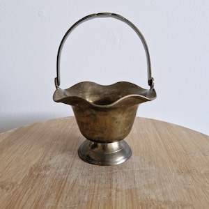May include: A small, antique brass basket with a scalloped rim and a curved handle. The basket sits on a pedestal base. The brass has a tarnished, aged appearance, suggesting it is vintage.