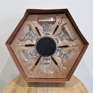 May include: A vintage cocktail set presented in a hexagonal box. The set comprises six clear glass cocktail glasses, six black-handled stirrers, and a central black coaster. The glasses feature a distinctive, rounded design. The box is brown.