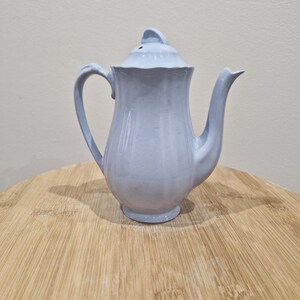 May include: A pale blue ceramic teapot with a lid, handle, and spout. The teapot is on a wooden surface. The teapot has a fluted design and a small knob on the lid. The teapot is a light blue colour.