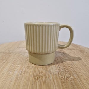 May include: A beige ceramic mug with vertical ribbed detailing on the upper section and a rounded handle. The mug sits on a wooden surface. The mug is designed for hot drinks.