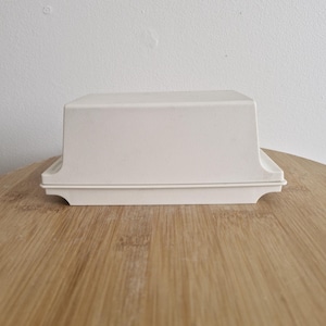 May include: A white rectangular butter dish with a lid. The dish has a slightly curved top and a decorative base. The butter dish is made of a solid, opaque material and sits on a wooden surface.