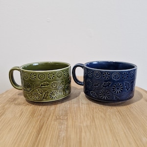 May include: Two ceramic mugs with handles, one in olive green and the other in navy blue. Both mugs feature a repeating floral and holly leaf pattern around the exterior. The mugs are sitting on a wooden surface.