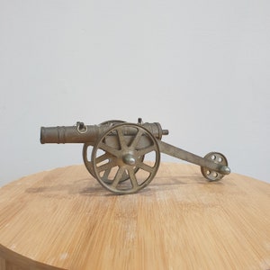 May include: A brass cannon figurine with a wheel and a barrel. The cannon is on a wooden surface.