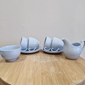 May include: A pale blue ceramic tea set comprising cups, saucers, a small bowl, and a creamer. The set is displayed on a round wooden table. The cups are stacked on saucers, and the creamer has a handle.