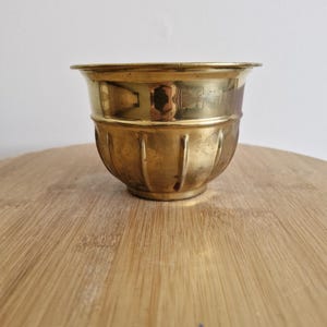May include: A small, antique brass bowl with a wide rim and a ribbed, rounded body. The bowl has a polished, reflective surface and sits on a wooden surface. The bowl is likely used for decorative purposes or as a small container.