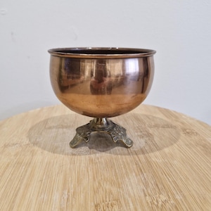 May include: A copper-coloured bowl with a rounded shape, set on a decorative, three-footed base. The bowl has a polished finish, reflecting the light. The base features an ornate, antique design. The bowl is likely used for decorative purposes.