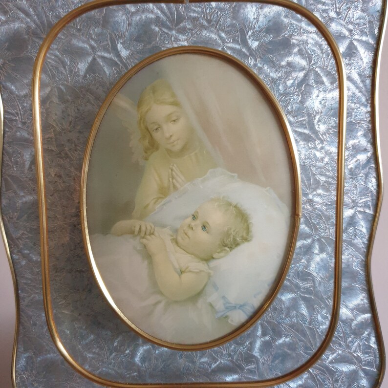 Op de afbeelding: Een vintage ovale fotolijst met een gouden rand en een lichtblauwe achtergrond. De lijst bevat een aquarel van een engel die over een slapende baby waakt.