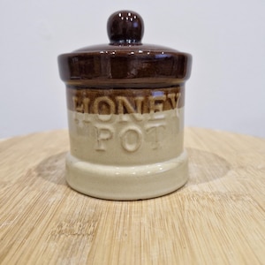 May include: A ceramic honey pot with a dark brown lid and a light beige base. The words "HONEY POT" are printed in a dark brown colour on the pot. The pot is sitting on a wooden surface.