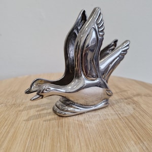 May include: A silver duck ornament, crafted with intricate detail. The wings are spread wide, suggesting flight. The polished metal reflects light, highlighting the form of the bird. This decorative item is likely intended for display, adding a touch of nature-inspired elegance.