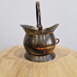 May include: A small, antique, brass bucket with a handle. The bucket has a rounded bottom and a slightly flared top. The bucket is tarnished and has a patina.