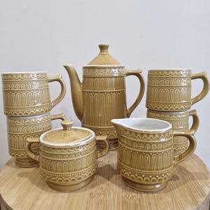 May include: A vintage tea set in a warm brown colour, featuring a teapot, creamer, sugar bowl with lid, and four mugs. The set has a repeating arched pattern and is displayed on a wooden surface. The teapot is approximately 20 cm tall.