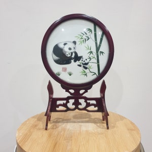 May include: A round, wooden framed embroidery depicting a black and white panda with two cubs on a bamboo branch. The embroidery is on a stand.