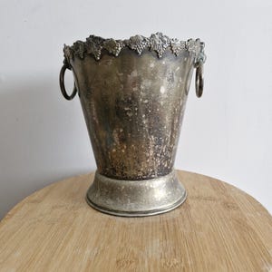 May include: A silver wine bucket with a decorative grape vine design around the rim. The bucket has two ring handles and a wide base. The metal has a tarnished appearance, giving it a vintage look. The bucket is sitting on a wooden surface.