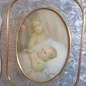 Op de afbeelding: Een vintage ovaal ingelijst beeld met een gouden lijst. De afbeelding toont een jong meisje met blond haar en een witte jurk die bidt voor een slapende baby met blond haar.