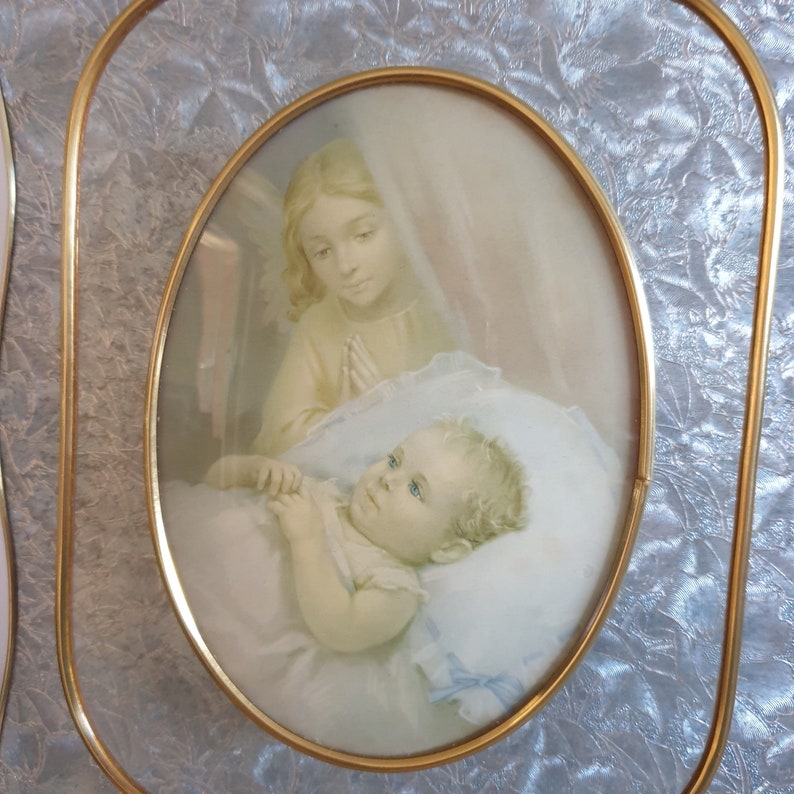 Op de afbeelding: Een vintage ovaal ingelijst beeld met een gouden lijst. De afbeelding toont een jong meisje met blond haar en een witte jurk die bidt voor een slapende baby met blond haar.