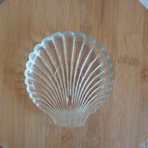 May include: A clear glass seashell dish with a ribbed texture. The dish is shaped like a scallop shell and is perfect for holding small items.