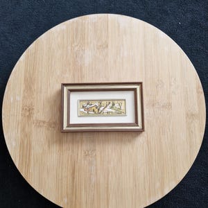 May include: A framed miniature landscape scene depicting a snow-covered village with trees. The artwork is set within a brown frame and rests on a light brown, circular wooden surface. The background is a dark blue fabric.