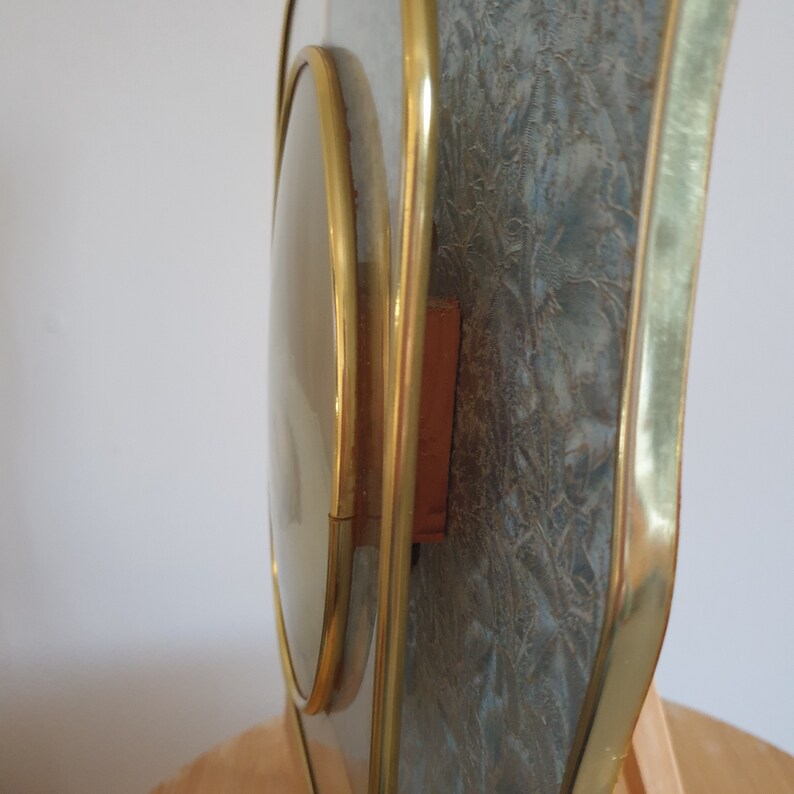Op de afbeelding: Een gouden geframed klok met een ronde, heldere wijzerplaat en een textuur, blauwe achtergrond. De klok staat op een houten oppervlak.