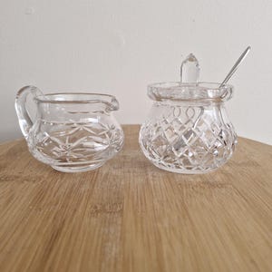May include: A clear, cut-glass creamer and sugar bowl set. The creamer has a handle and spout. The sugar bowl has a lid and a small spoon. Both pieces have a diamond pattern and sit on a wooden surface.