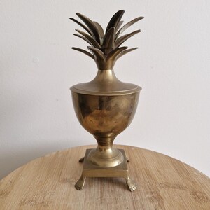 May include: A vintage brass pineapple-shaped container with a lid. The container has a pedestal base with small feet. The pineapple top is detailed with individual leaves. The container is likely used for decorative purposes.