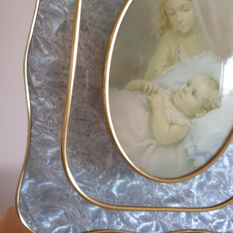Op de afbeelding: Een vintage ovale fotolijst met een gouden rand en een lichtblauwe, textuurrijke achtergrond. De lijst bevat een foto van een vrouw die een slapende baby vasthoudt.