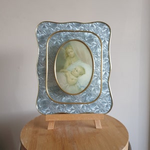 Op de afbeelding: Een vintage zilverkleurig fotolijstje met een gouden rand en een gegolfde rand. Het lijstje bevat een afbeelding van een engel die een baby vasthoudt. Het lijstje staat op een houten ezel.