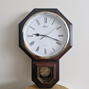 May include: An octagonal wall clock with a dark wood frame and a gold-toned bezel. The white clock face features black Roman numerals, the text "Flenders" and "QUARTZ," and black hands. A lower glass panel displays a gold pendulum and "REGULATOR."