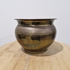 May include: A small, round, hammered brass bowl with a wide, flared rim. The metal surface features a textured, dimpled pattern across the exterior, displaying a mix of dark, tarnished tones and bright, polished gold-coloured metallic highlights.
