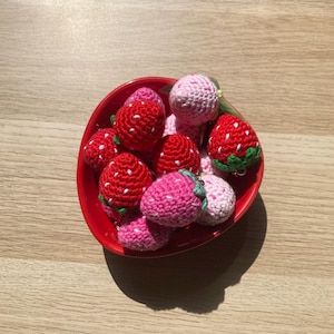 Peut inclure: Un bol rouge rempli de plusieurs fraises crochetées. Les fraises sont de différentes nuances de rouge et de rose, avec des détails de graines blanches et des feuilles vertes. Le bol est posé sur une surface en bois brun clair.