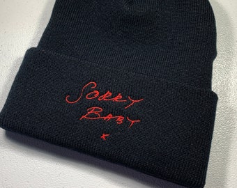 May include: Black beanie with a red embroidered design that says "Sorry Baby" with a small red star below the text.