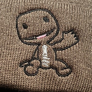 K&ouml;nnte beinhalten: Braune Strickm&uuml;tze mit einem schwarzen gestickten Comic-Charakter mit rosa Mund und wei&szlig;en Augen.