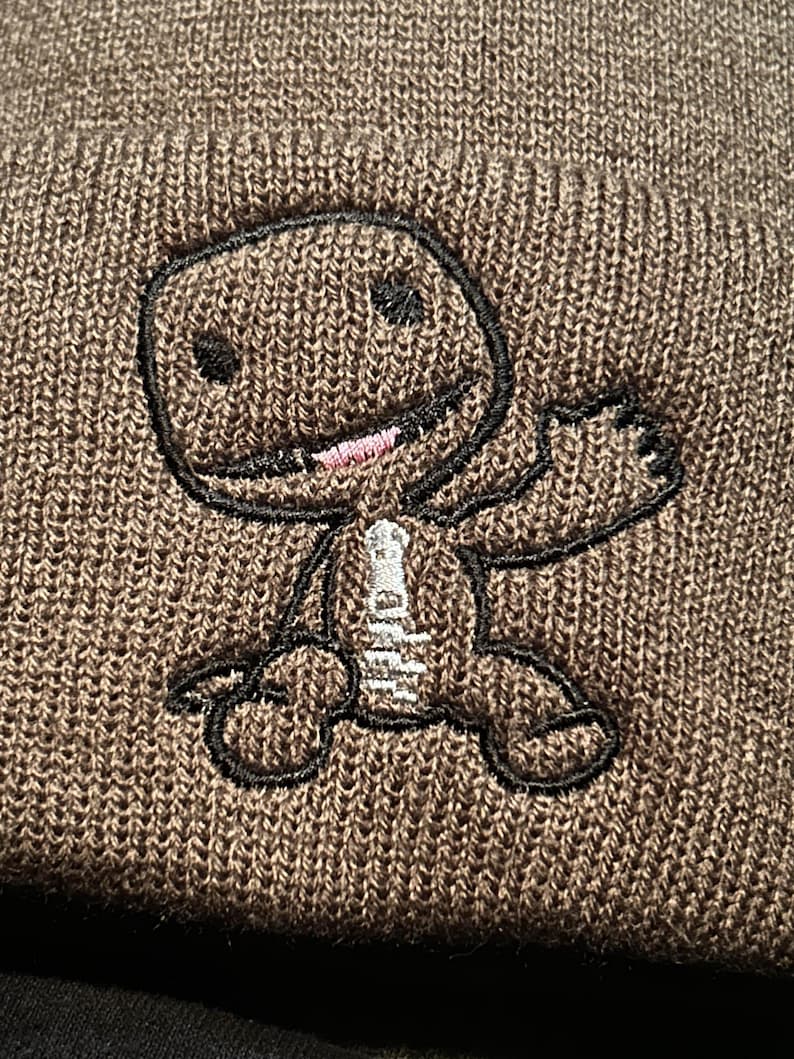 K&ouml;nnte beinhalten: Braune Strickm&uuml;tze mit einem schwarzen gestickten Comic-Charakter mit rosa Mund und wei&szlig;en Augen.