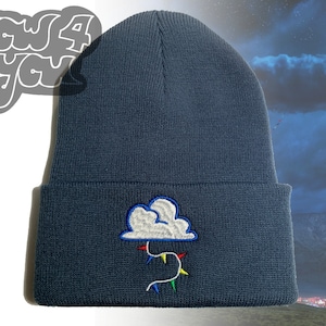 Pode incluir: Um gorro de malha azul escuro com uma nuvem bordada branca e luzes de corda coloridas penduradas nele.