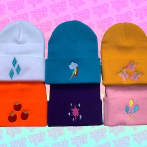 Op de afbeelding: Zes kleurrijke gebreide mutsen met geborduurde My Little Pony-figuren. De mutsen zijn wit, turquoise, geel, oranje, paars en roze. De figuren zijn Diamond Tiara, Rainbow Dash, Fluttershy, Applejack, Twilight Sparkle en Pinkie Pie.
