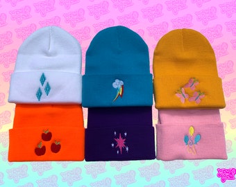 Pode incluir: Seis gorros de malha coloridos com personagens My Little Pony bordados. Os gorros são brancos, turquesa, amarelos, laranja, roxos e rosa. Os personagens são Diamond Tiara, Rainbow Dash, Fluttershy, Applejack, Twilight Sparkle e Pinkie Pie.