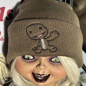 Puede incluir: Un gorro de punto marrón con un personaje de dibujos animados bordado en negro y blanco en la parte delantera. El personaje está saludando con un brazo y tiene una cara sonriente.