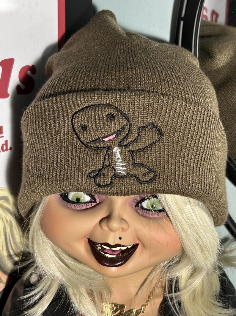 K&ouml;nnte beinhalten: Eine braune Strickm&uuml;tze mit einem schwarzen und wei&szlig;en gestickten Comic-Charakter auf der Vorderseite. Der Charakter winkt mit einem Arm und hat ein l&auml;chelndes Gesicht.