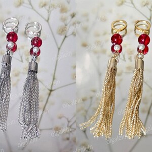 May include: Dangle earrings with chain tassels, one pair silver, the other gold. Each earring has red and white beads, and a ring. The earrings are shown on a reflective surface with a floral background.