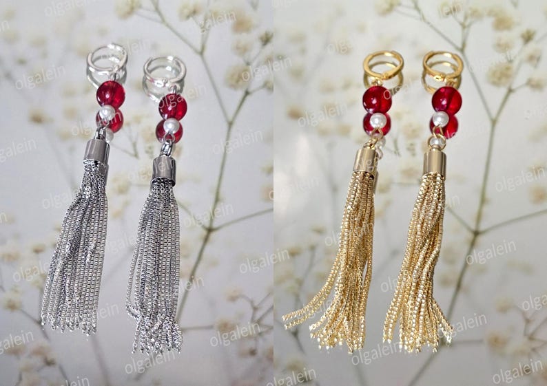 May include: Dangle earrings with chain tassels, one pair silver, the other gold. Each earring has red and white beads, and a ring. The earrings are shown on a reflective surface with a floral background.