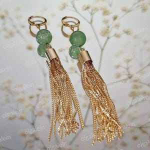 May include: A pair of gold-toned tassel earrings with green gemstone accents. The earrings feature a gold-colored hoop and a cylindrical top, with long, dangling chains. The background is a blurred, light-colored floral pattern.