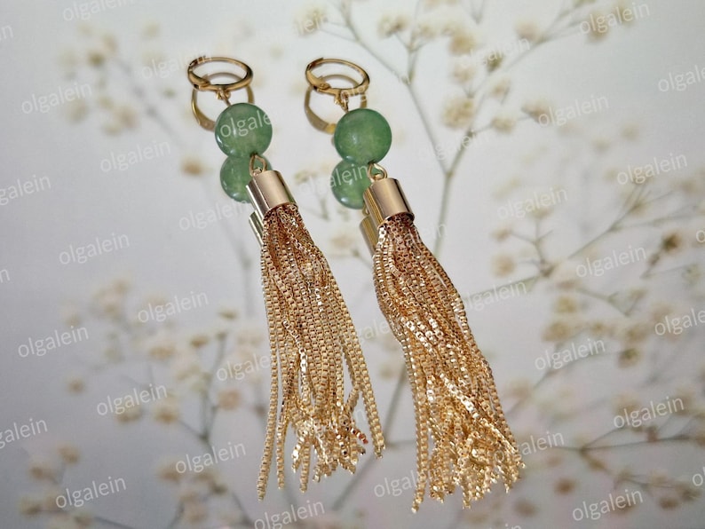 May include: A pair of gold-toned tassel earrings with green gemstone accents. The earrings feature a gold-colored hoop and a cylindrical top, with long, dangling chains. The background is a blurred, light-colored floral pattern.