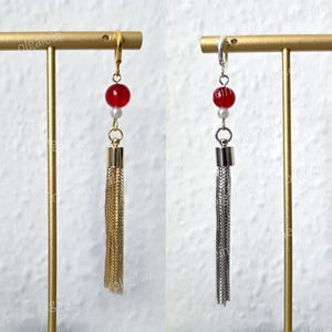 May include: Earrings featuring a red bead, a small white bead, and a chain tassel. One pair has gold-coloured metal, and the other has silver-coloured metal. The earrings are displayed on a gold-coloured stand.